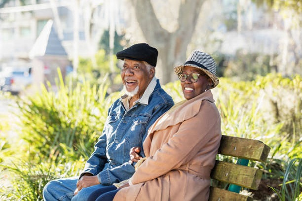 A senior African-American couple sitting together on a park bench on a sunny autumn day. They are side by side, arm in arm, relaxing, smiling. The man is in his 70s and the woman is in her 60s.