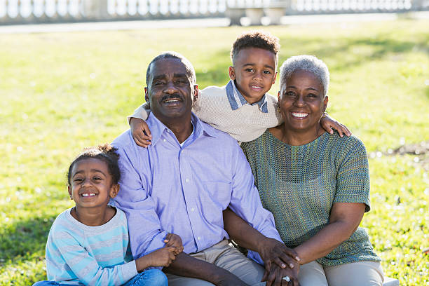 Portrait of an African American multigeneration family.  Two cute children are in the park with their grandparents, sitting on a bench.  The little boy is standing behind, with his arms around his grandparents' shoulders.